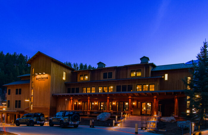 The exterior of Sundance Resort at dusk, a large wood-sided lodge glowing with warm lights, surrounded by pine trees and mountains
