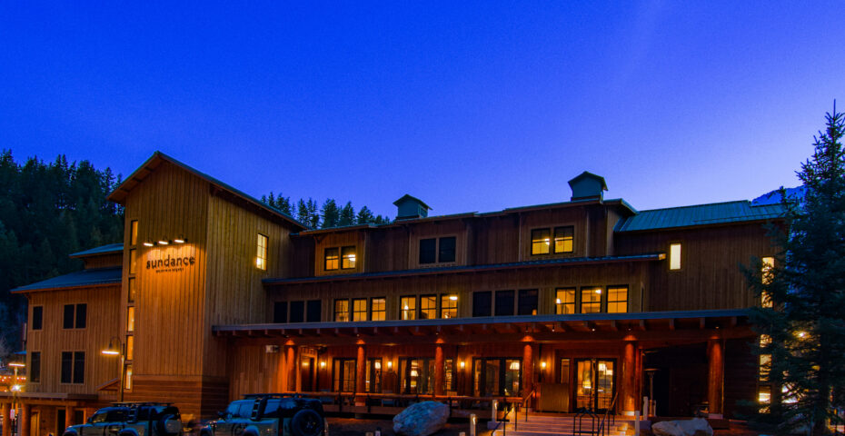 The exterior of Sundance Resort at dusk, a large wood-sided lodge glowing with warm lights, surrounded by pine trees and mountains